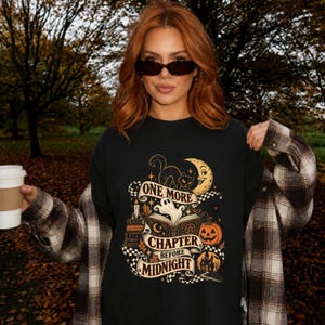 Può includere: Una t-shirt nera a girocollo con una stampa grafica raffigurante un fantasma che emerge da un libro aperto, un gatto nero, una luna crescente, una zucca e una casa stregata. Il testo recita One More Chapter Before Midnight.