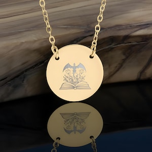 Puede incluir: Un collar con colgante dorado con un dije circular. El dije presenta un diseño grabado de un libro abierto con flores y una figura alada. La cadena del collar también es dorada.