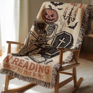 A Halloween-themed blanket with a skeleton reading a book and a jack-o-lantern is draped over a wooden rocking chair.