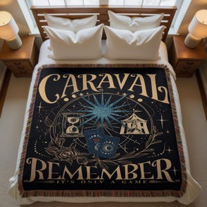 May include: Black throw blanket with a gold and blue design. The blanket features the word "CARAVAL" at the top and "REMEMBER" at the bottom, with a central illustration of a starburst, hourglass, and tarot cards. The blanket has a fringed edge.