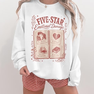 May include: White sweatshirt with a red graphic that reads "FIVE-STAR Emotional Damage." The graphic is designed to look like an open book with illustrations and text. The person is wearing red and white checkered shorts.