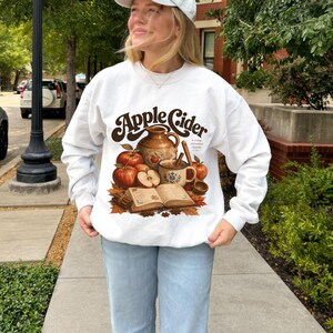 White sweatshirt with "Apple Cider" text and fall graphic of apples, mug, and book.