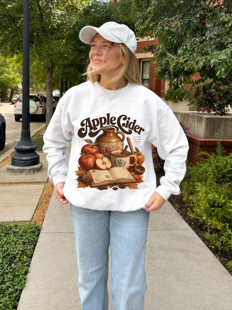 White sweatshirt with "Apple Cider" text and fall graphic of apples, mug, and book.
