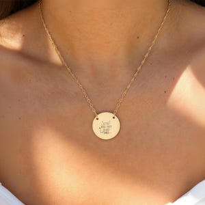 A gold-colored necklace with a round pendant featuring a message engraved on it.