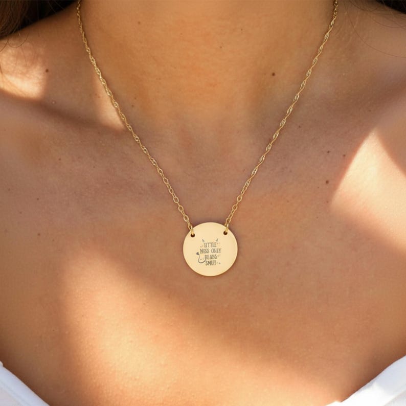 A gold-colored necklace with a round pendant featuring a message engraved on it.