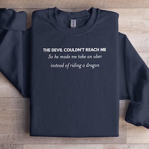 Puede incluir: Sudadera azul marino con el texto blanco: "THE DEVIL COULDN'T REACH ME So he made me take an uber instead of riding a dragon". La prenda está doblada, mostrando las mangas.
