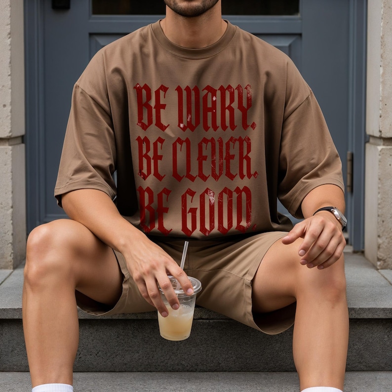 Man wearing a brown t-shirt with red text that reads "BE WARY. BE CLEVER. BE GOOD."
