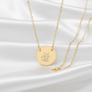 A gold-colored necklace with a round pendant featuring a message engraved on it.