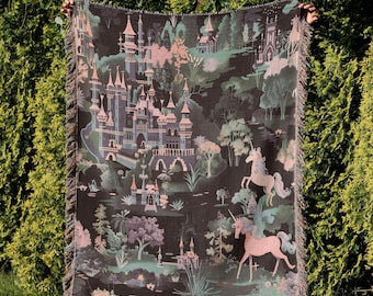 May include: A dark blanket with a whimsical castle and unicorn design. The blanket features a detailed illustration in shades of black, pink, and green, with a fringed edge. The blanket is held up in front of a green hedge.