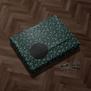 A green and gold patterned gift box with a black circular design on the front, placed on a wooden floor.