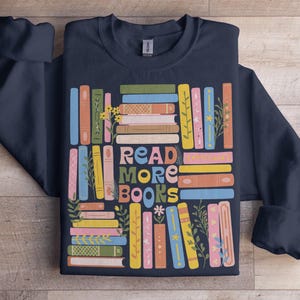 Puede incluir: Sudadera azul marino con un gráfico colorido de libros apilados y la frase "READ MORE BOOKS" en una fuente divertida. Los libros son de varios colores, con detalles florales.