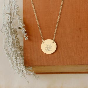 A gold-colored necklace with a round pendant featuring a message engraved on it, resting on a wooden surface.