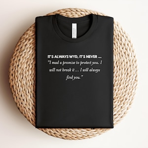 May include: Black t-shirt with the text "IT'S ALWAYS WYD, IT'S NEVER..." and a quote in white. The shirt is folded and placed on a round, woven, beige placemat. The shirt has a crew neck.