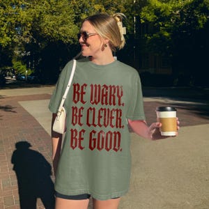 Woman in olive green t-shirt with "BE WARY. BE CLEVER. BE GOOD." text.