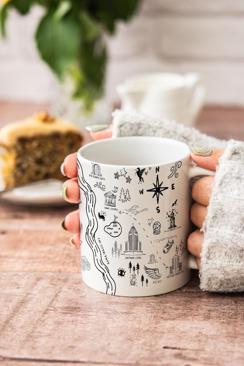 A person's hands holding a white mug with various black and white illustrations and text on it, placed on a wooden surface.