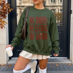Green sweatshirt with "Be Wary. Be Clever. Be Good." text, worn by a woman outside.