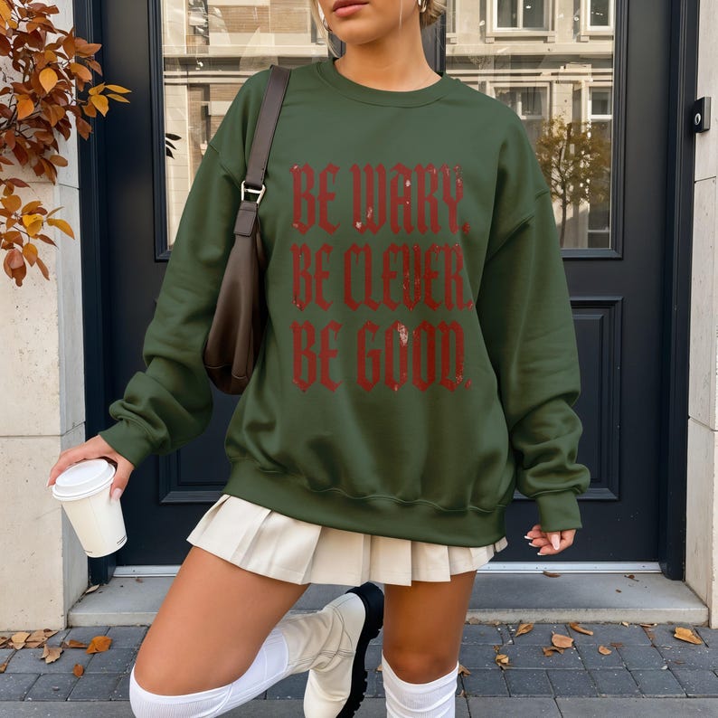 Green sweatshirt with "Be Wary. Be Clever. Be Good." text, worn by a woman outside.