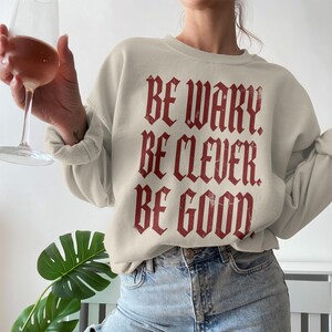 Cream sweatshirt with red gothic text "BE WARY BE CLEVER BE GOOD".