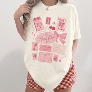 Cream t-shirt with pink "The Book Club" graphic featuring stamps and playing cards.