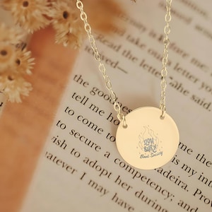 May include: A gold-colored necklace with a circular pendant. The pendant has the text "Slow Burn Book Society" and a flame graphic. The necklace is on a chain and rests on a book page.