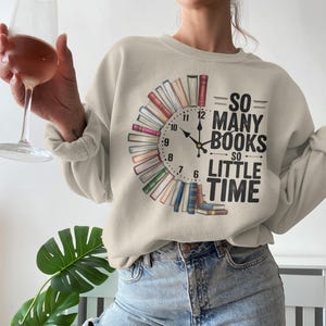 Op de afbeelding: Beige sweatshirt met een klokontwerp gemaakt van boeken. De klokwijzers wijzen naar 12:00 uur. De tekst luidt "So Many Books So Little Time". Een casual, comfortabele stijl.