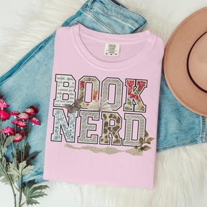 Pink t-shirt with "BOOK NERD" text in distressed, patterned letters.