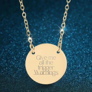 May include: Gold-colored necklace with a circular pendant. The pendant has the phrase "Give me all the trigger Warnings" engraved in a sans-serif font. The chain is a delicate link style, and the background is a dark teal.