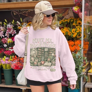Pink crewneck sweatshirt with "Meet Me at the Bookstore" graphic and book-themed illustration.