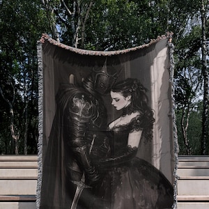 Op de afbeelding: Een zwart-wit geweven sprei met een romantische illustratie van een ridder in harnas die een vrouw in een gotische jurk omhelst. De deken heeft een gefranjerde rand en wordt buiten getoond.