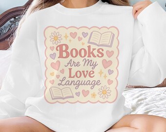 May include: White sweatshirt with a graphic that reads "Books Are My Love Language." The design features hearts, flowers, and open books in soft pink and peach tones. A cozy, book-themed apparel item.