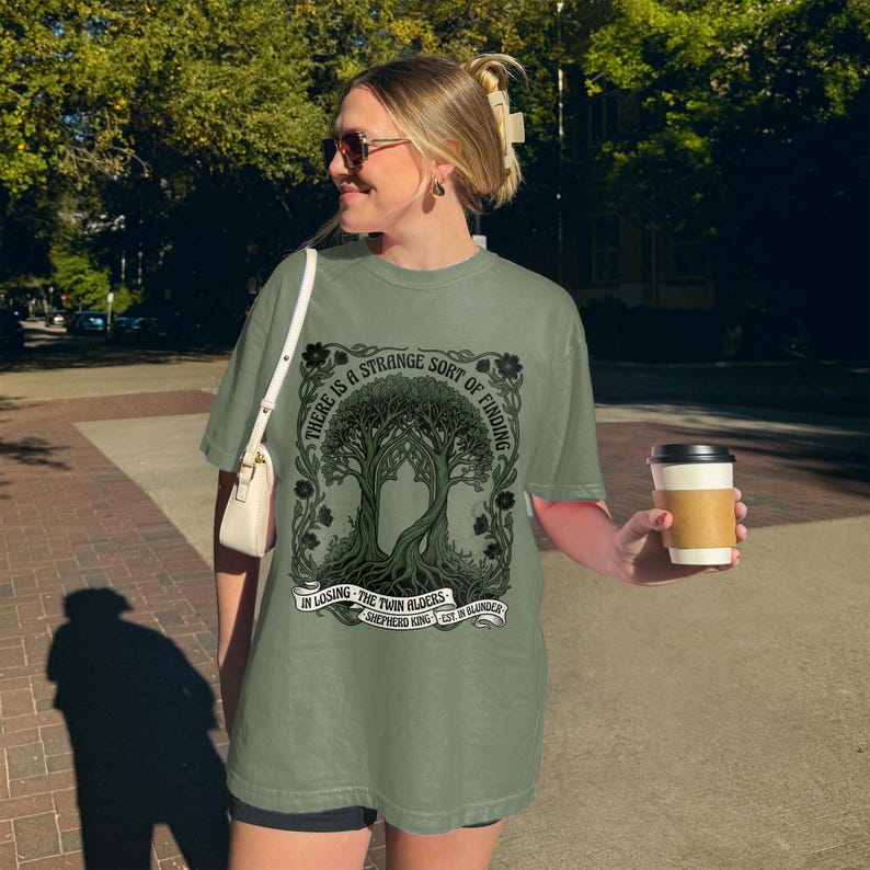 T-shirt with a tree design and text: "There is a strange sort of finding in losing the twin alders. Shepherd King Est. in Blunder."