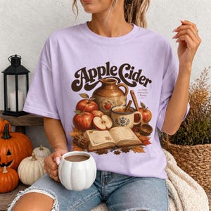 T-shirt graphic with "Apple Cider" text, jug, apples, mug, and open book.