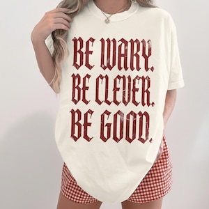 Cream t-shirt with red gothic text reading "Be Wary. Be Clever. Be Good."