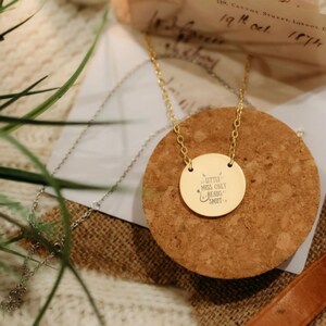 A gold-colored necklace with a round pendant featuring a message engraved on it, resting on a cork coaster.