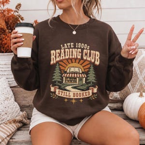 Brown sweatshirt with "Late 1900s Reading Club Still Booked" text and bookstore illustration.