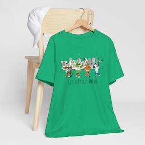 Könnte beinhalten: Ein grünes T-Shirt mit einer Grafik einer Gruppe von Personen vor einer Stadtkulisse. Der Text "It's a Philly Thing" ist unter der Grafik gedruckt.