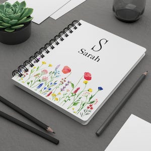 May include: A white spiral-bound notebook featuring a watercolor illustration of colorful wildflowers along the bottom edge. The cover displays the letter S and the name Sarah in black serif font centered in the upper portion of the notebook.