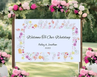 Op de afbeelding: Een welkomstbord voor een bruiloft met de tekst "Welcome To Our Wedding" en de namen Ashley & Jonathan, met de datum 05.28.25. Het bord is versierd met bloemstukken in roze en wit, en is omgeven door meer bloemstukken.