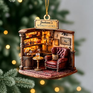 Puede incluir: Un adorno colgante en miniatura que representa un salón de whisky con un sillón capitoné de color rojo oscuro, una mesa redonda pequeña con un vaso de líquido ámbar y estantes llenos de botellas. Un letrero dorado arriba dice Jackson's WHISKEY LOUNGE.