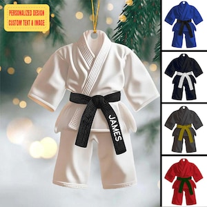 May include: White karate uniform ornament with a black belt, personalised with the name "JAMES". Additional ornaments in blue, black, and red with different coloured belts are also shown. The ornament has a gold hanging string and text that reads "PERSONALIZED DESIGN CUSTOM TEXT & IMAGE".