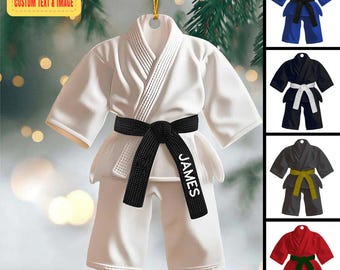 May include: White karate uniform ornament with a black belt, personalized with the name "JAMES". Additional ornaments in blue, black, and red with different colored belts are also shown. The ornament has a gold hanging string and text that reads "PERSONALIZED DESIGN CUSTOM TEXT & IMAGE".