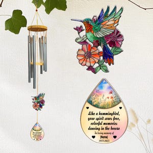Puede incluir: Una colección de elementos decorativos con temática de colibríes. Incluye una carillón de viento con tubos plateados y una parte superior de madera, un colibrí de vidriera con flores y un recuerdo en forma de lágrima con texto.