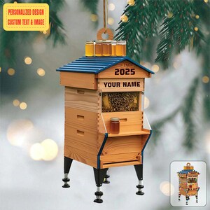 Könnte beinhalten: Ein hölzernes Bienenwabenornament mit blauem Dach und schwarzen Beinen. Das Ornament hat ein klares Fenster, das das Innere des Bienenstocks zeigt. Der Text "2025" und "YOUR NAME" sind auf der Vorderseite aufgedruckt. Kleine Gläser Honig stehen oben.