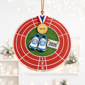 Op de afbeelding: Een rond acryl ornament met een rood hardloopbaandesign, witte baanmarkeringen en de cijfers 1, 2 en 3. In het midden staan blauwe en witte sportschoenen, een gouden medaille met de tekst ALEX en een wit bordje met het jaartal 2026.