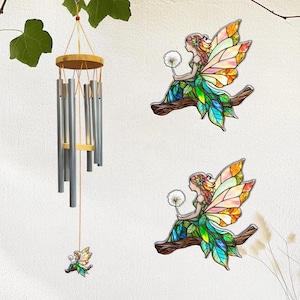 Puede incluir: Un carillón de viento con tubos plateados y una parte superior de madera cuelga cerca de dos decoraciones de hadas de vidrieras. Las hadas están representadas con alas y vestidos coloridos, sosteniendo dientes de león mientras están posadas en ramas. El carillón tiene un hada pequeña en la parte inferior.