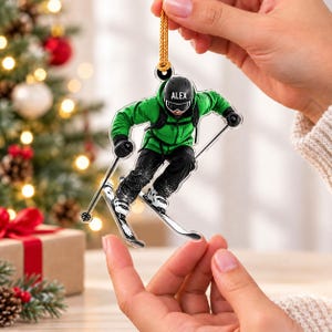 Puede incluir: Un adorno navideño de acrílico transparente que muestra a un esquiador con chaqueta verde brillante, pantalones negros y un casco negro con el nombre ALEX impreso en el frente. El esquiador está en posición de descenso con bastones y esquís.