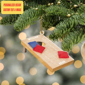 Könnte beinhalten: Holz-Cornhole-Spiel-Ornament mit roten und blauen Beanbags. Das Ornament trägt den Namen "WILLIAM" und das Jahr "2025". Der Schmuck hängt an einer goldenen Kette an einem Weihnachtsbaum.