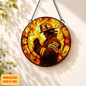 Puede incluir: Vidriera redonda que representa a un bombero de perfil, sobre un fondo de llamas. El bombero lleva un uniforme marrón con rayas amarillas y el nombre "DAVID" y el número "373". Cuelga de una cadena negra.