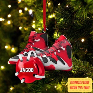 May include: A red and black wrestling shoe ornament with white accents and a matching red headgear ornament. The headgear has the name "JACOB" in white. The ornament hangs from a red ribbon against a blurred Christmas tree background.