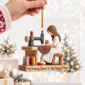 May include: A decorative hanging ornament featuring a sculpted figure with brown hair in a white dress sitting at a wooden sewing table. The table displays the name Sophia, a black sewing machine, colorful thread spools, and the text My Sewing Space Is My Happy Place.
